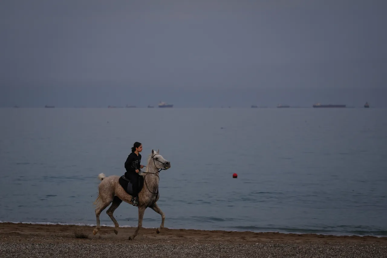A rider gallops along a beach as oil tankers line up in the Strait of Hormuz near Khor Fakkan, UAE, March 11, 2026. (Photo/Altaf Qadri)